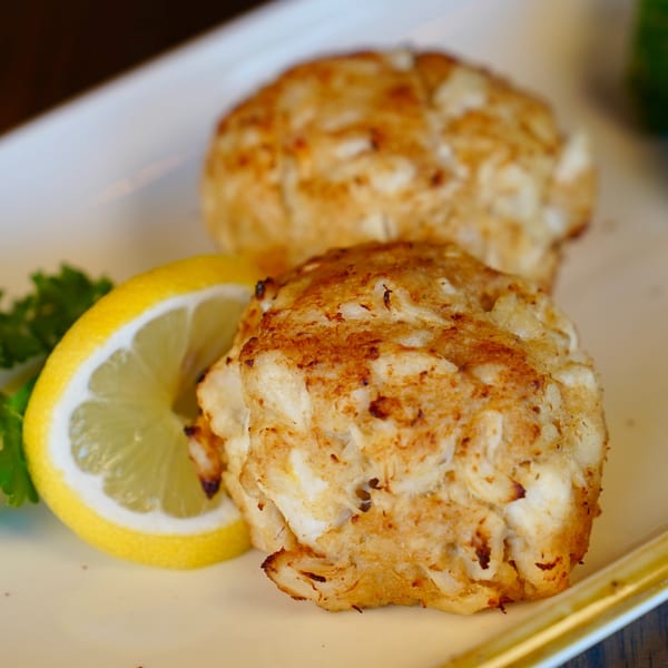 Two golden-brown crab cakes with a lemon wedge and parsley on a white plate at Colonial Bar & Grill.