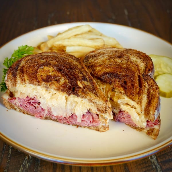 Colonial Bar & Grill's Reuben sandwich, cut in half, with fries, parsley, and pickles on a white plate.