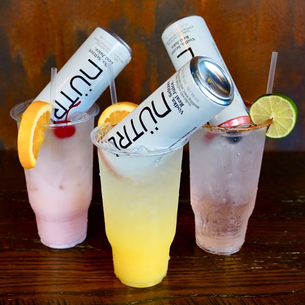 Three NÜTRL vodka seltzers in plastic cups with fruit garnishes on a wooden table.