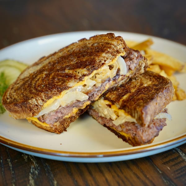 Colonial Bar & Grill's patty melt, cut in half, with fries and pickles on a white plate.
